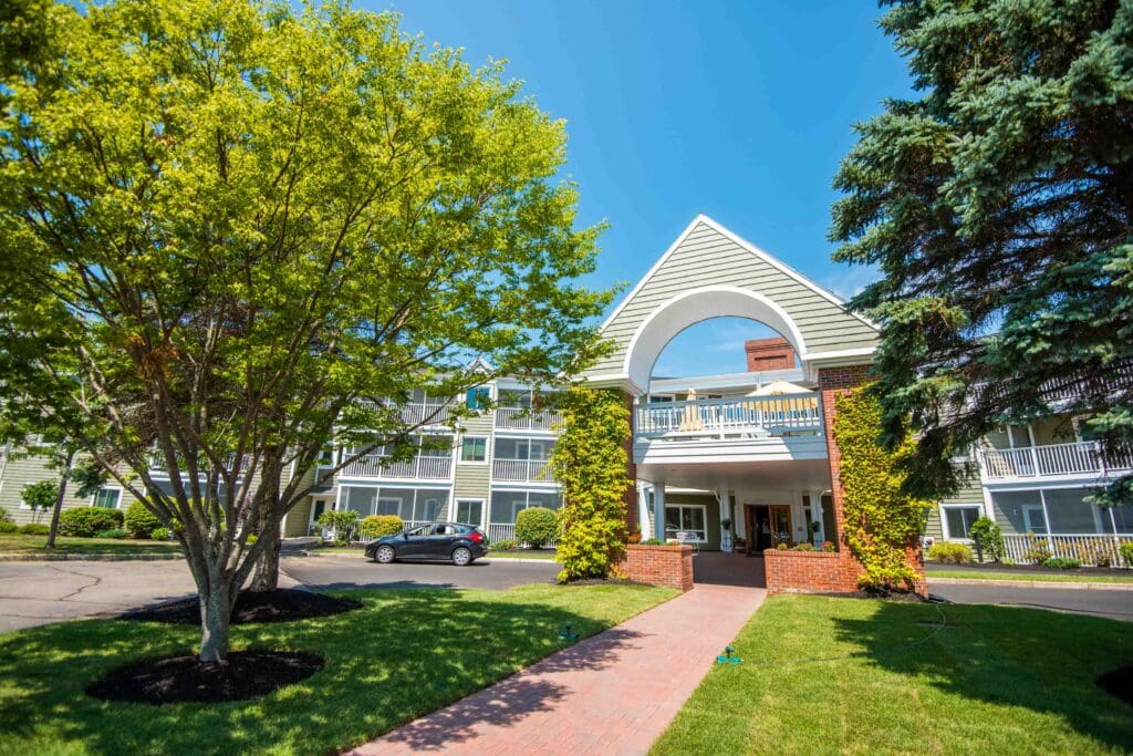 The Highlands senior living community main building during the day, in front of a walking path, green trees, and grass.